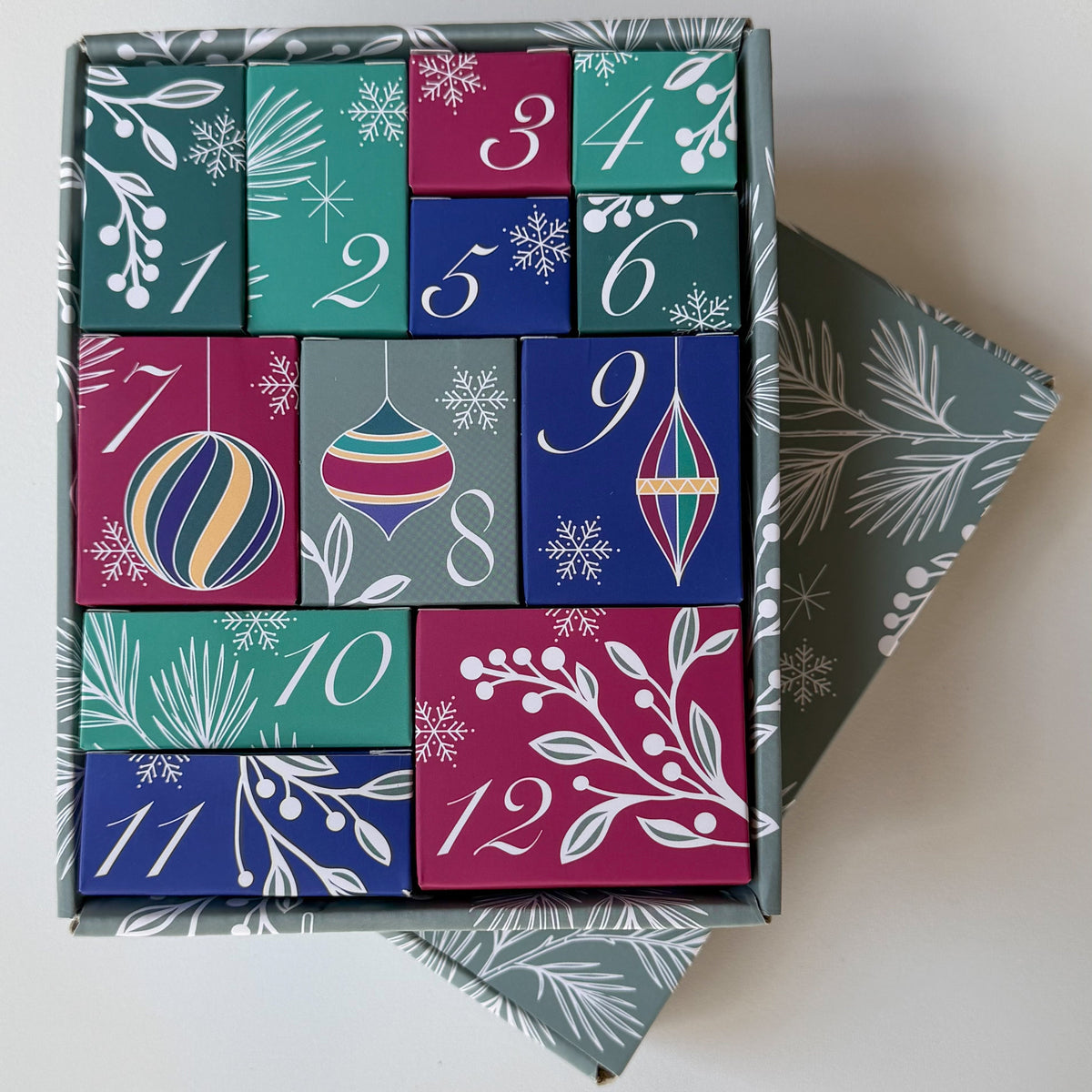 An open 12-box advent calendar box with colorful numbered compartments decorated with festive Christmas illustrations, shown on a light neutral background. Customized by Style Dots, available in gold and silver, launched October 2026