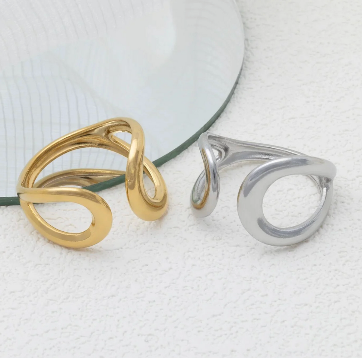 Two rings in silver and gold, that have a distinctive twisted wrap-style ring on a textured white surface, with a curved clear glass prop in the background.