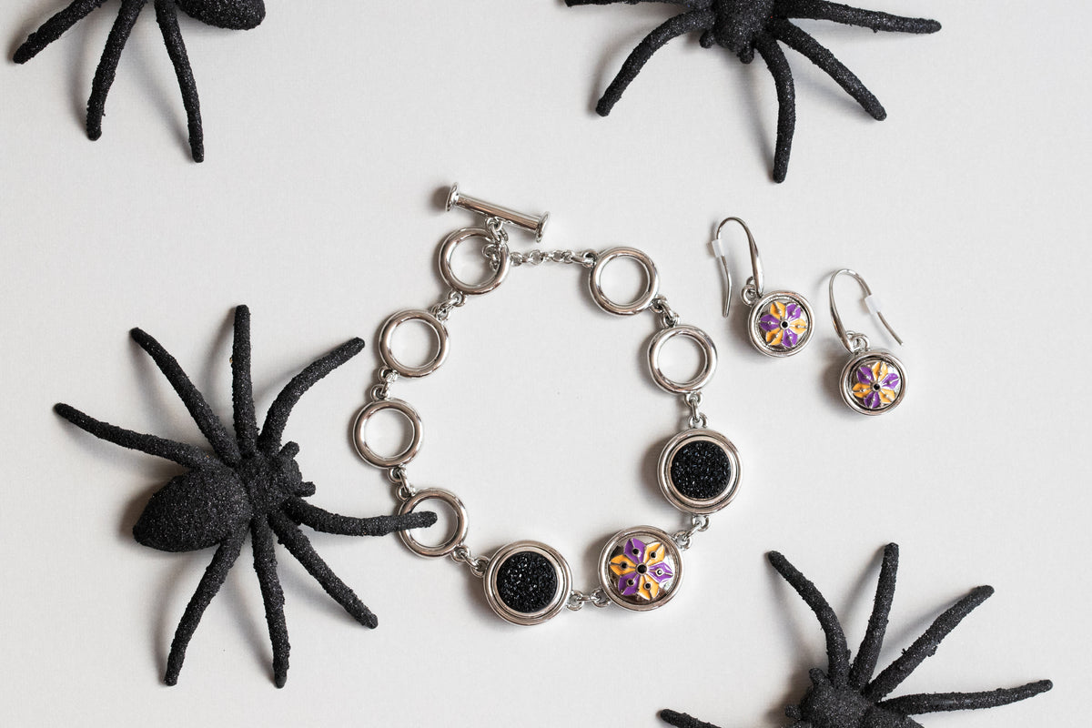 Silver-toned jewelry set featuring a bracelet and matching earrings with circular links, one black textured disc, and one purple-and-gold iridescent disc, arranged on a white background with black spider decorations. Release is for Halloween 2026.