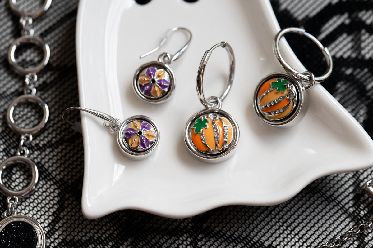 Silver-toned jewelry set including 2 sets of different drop earrings, displayed on a white scalloped dish over a black patterned fabric. The charms feature floral and pumpkin designs for Halloween 2026