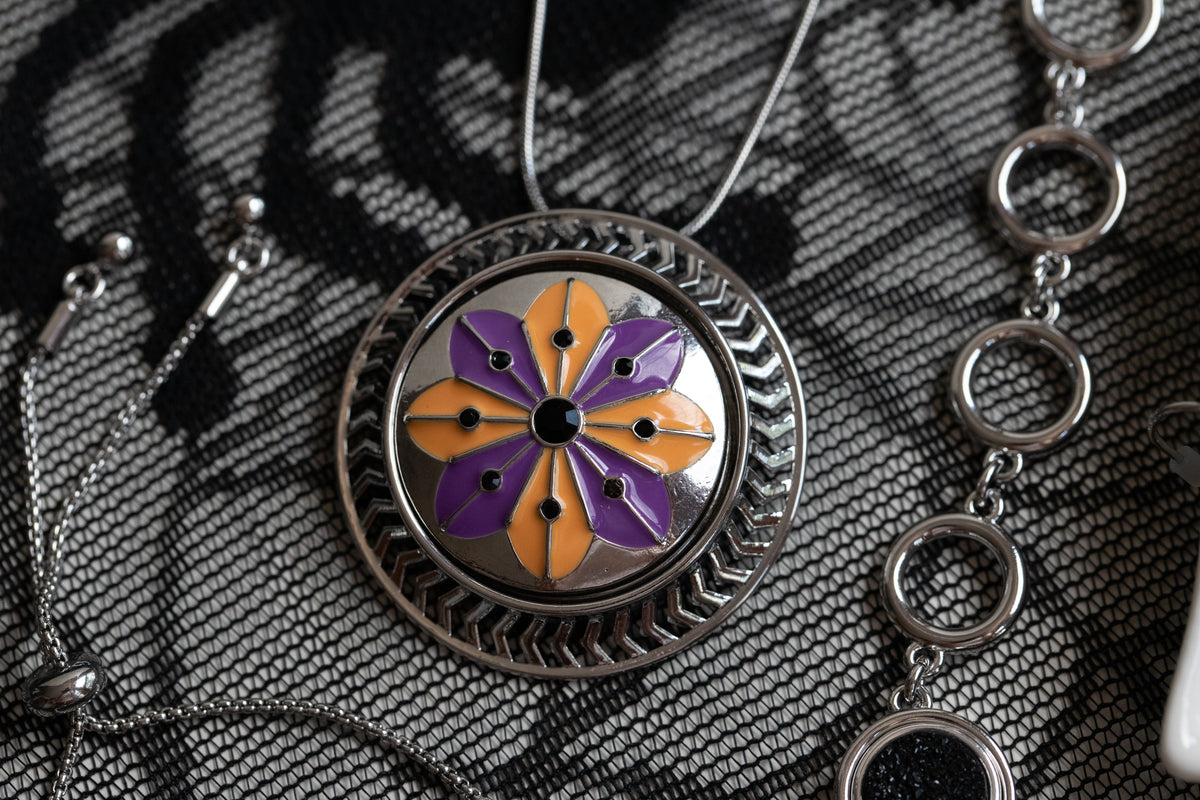 A close-up of a round silver pendant necklace with a floral Statement Dot design in purple and orange enamel, resting on a black and white patterned fabric alongside other silver jewelry.