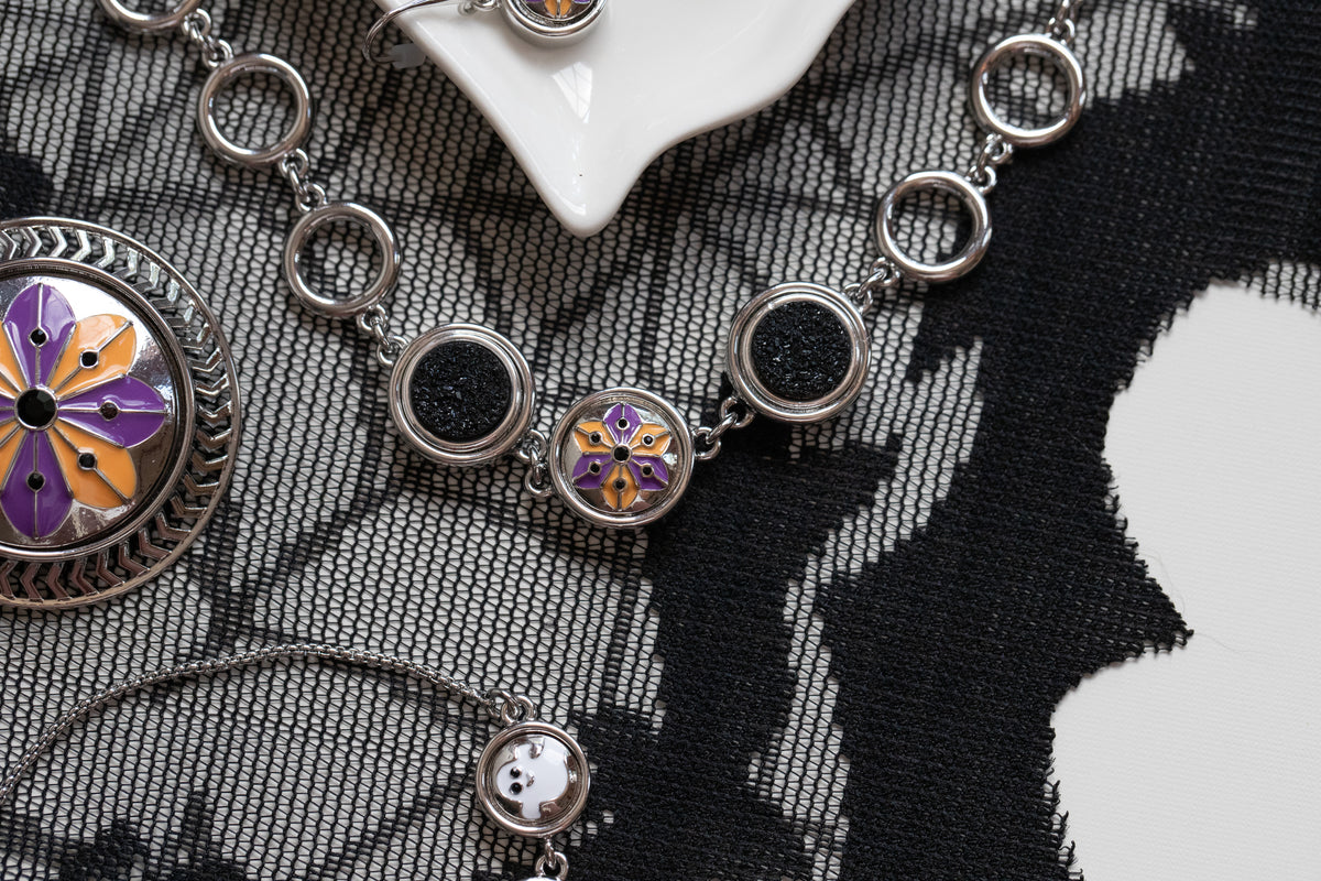 Silver-toned jewelry pieces with circular links and floral enamel details in purple, orange, and yellow, arranged on a black-and-white laces patterned fabric background for Halloween 2026