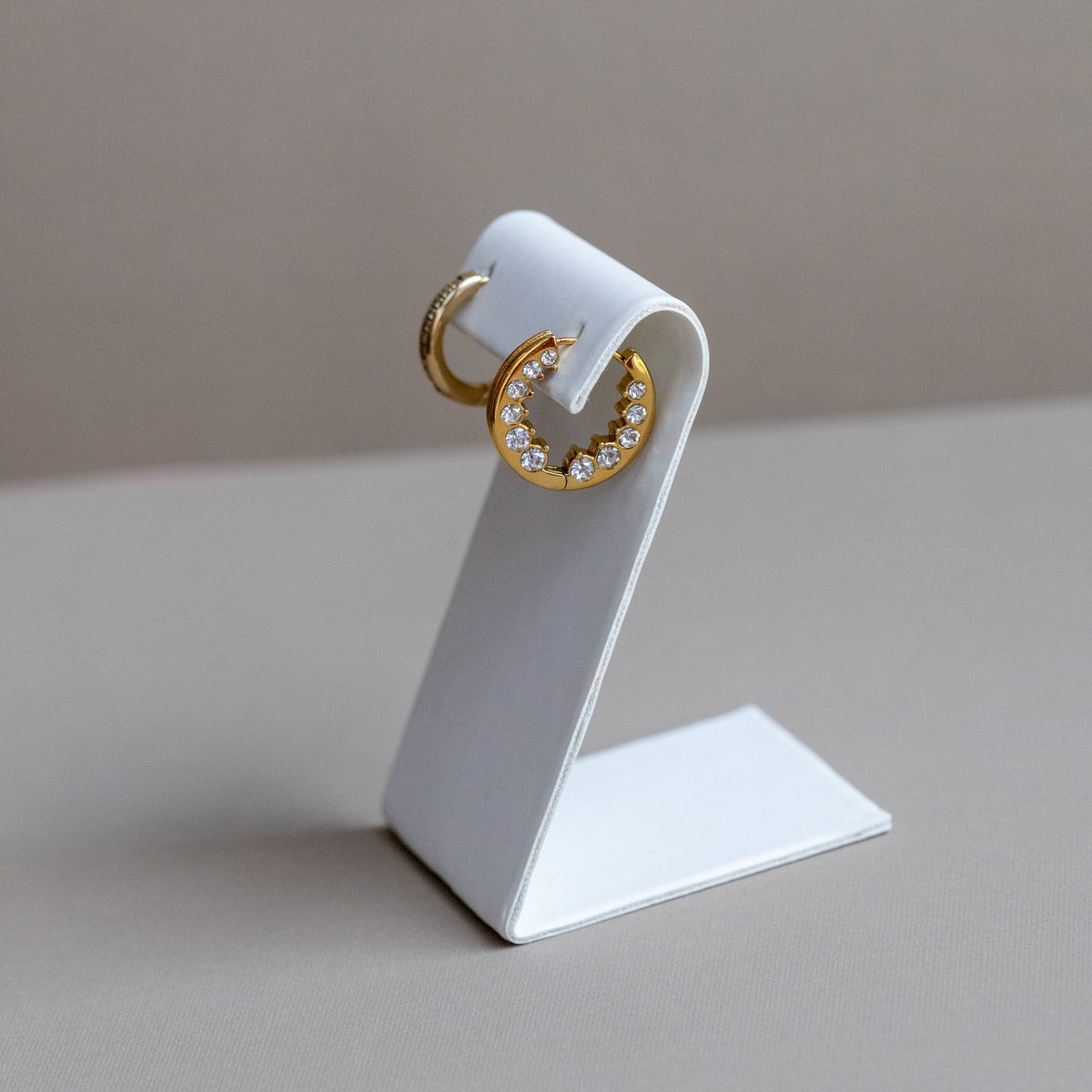 A pair of gold hoop earrings, one plain and one set with small clear stones, displayed on a white stand against a neutral beige background.