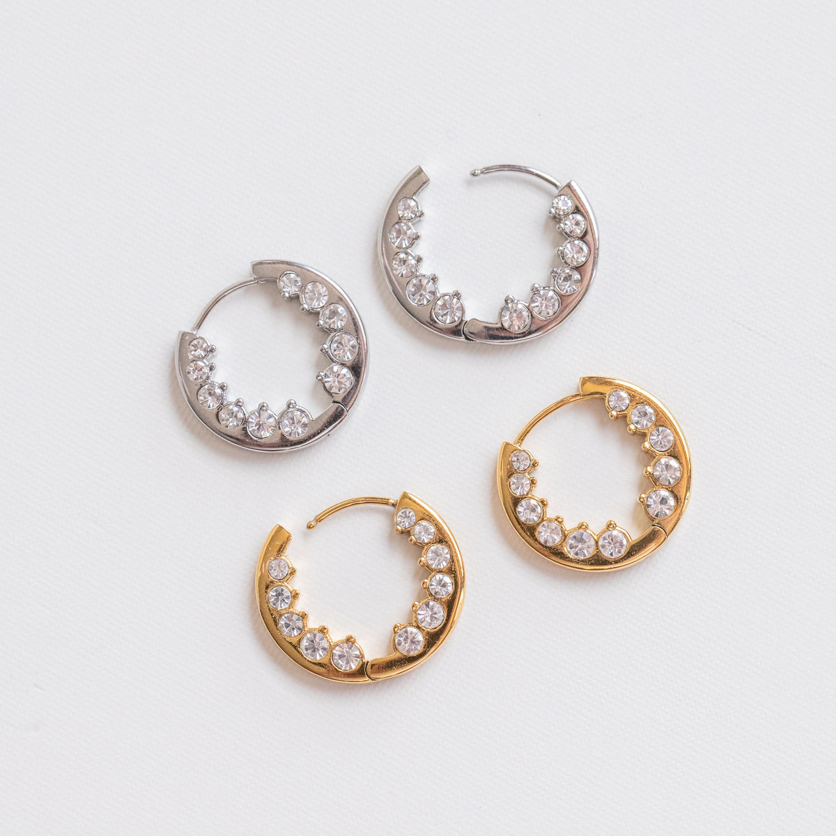 Four crescent-shaped hoop earrings with clear stones, shown in silver and gold finishes on a light gray textured background.