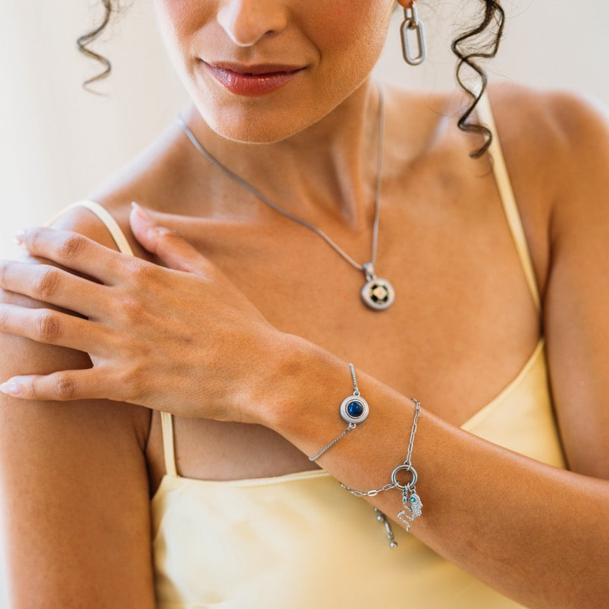 A close-up lifestyle shot of a woman wearing a silver-toned jewelry set, including a delicate necklace with a circular medallion pendant, a matching bracelet with a blue stone, and a charm, while wearing a pale yellow camisole.