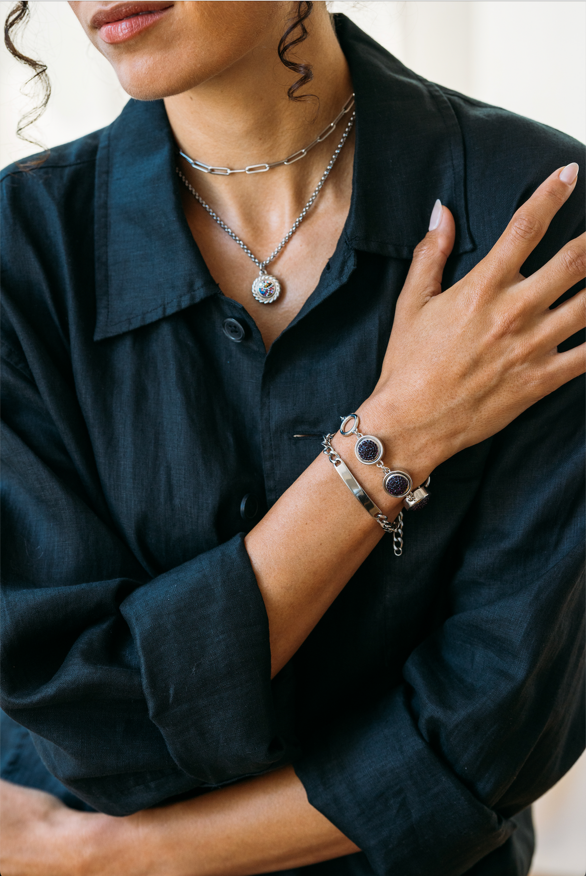 A close-up lifestyle shot of a model wearing a silver-toned layered jewelry set, including a pendant necklace, chain necklace, bracelet, and bangle, styled with a dark linen button-up shirt.