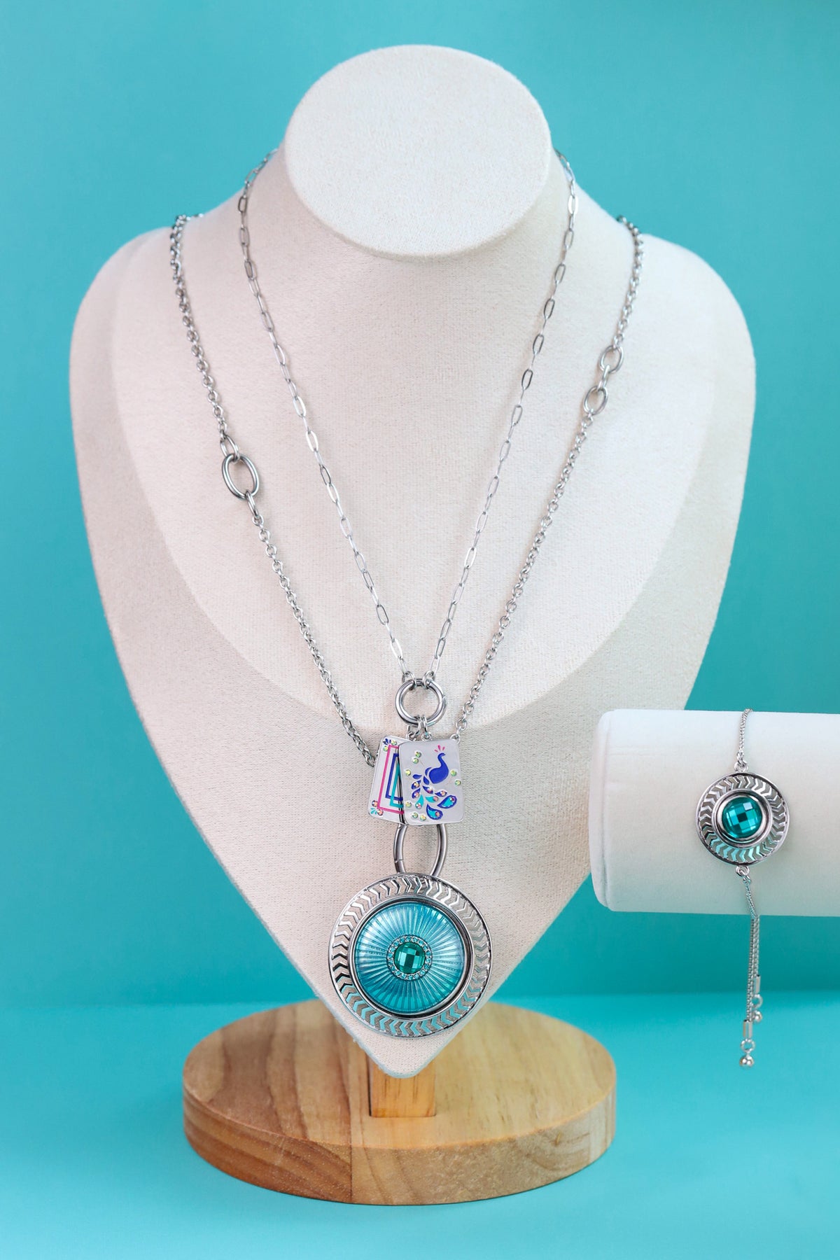 Silver jewelry set displayed on a white mannequin bust against a bright turquoise background, featuring a layered necklace with a round turquoise-blue and silver pendant, a matching bollo bracelet on a white stand, and a small matching Dotlet.