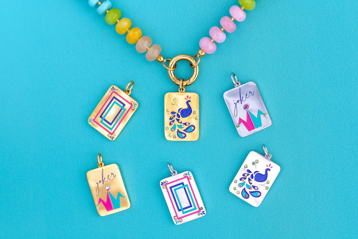 A beaded charm keeper necklace with a gold clasp and six colorful rectangular pendant charms featuring peacock and crown designs, arranged on a bright blue background.  The jewelry collection is inspired by Eastern Asian Mahjong tiles.