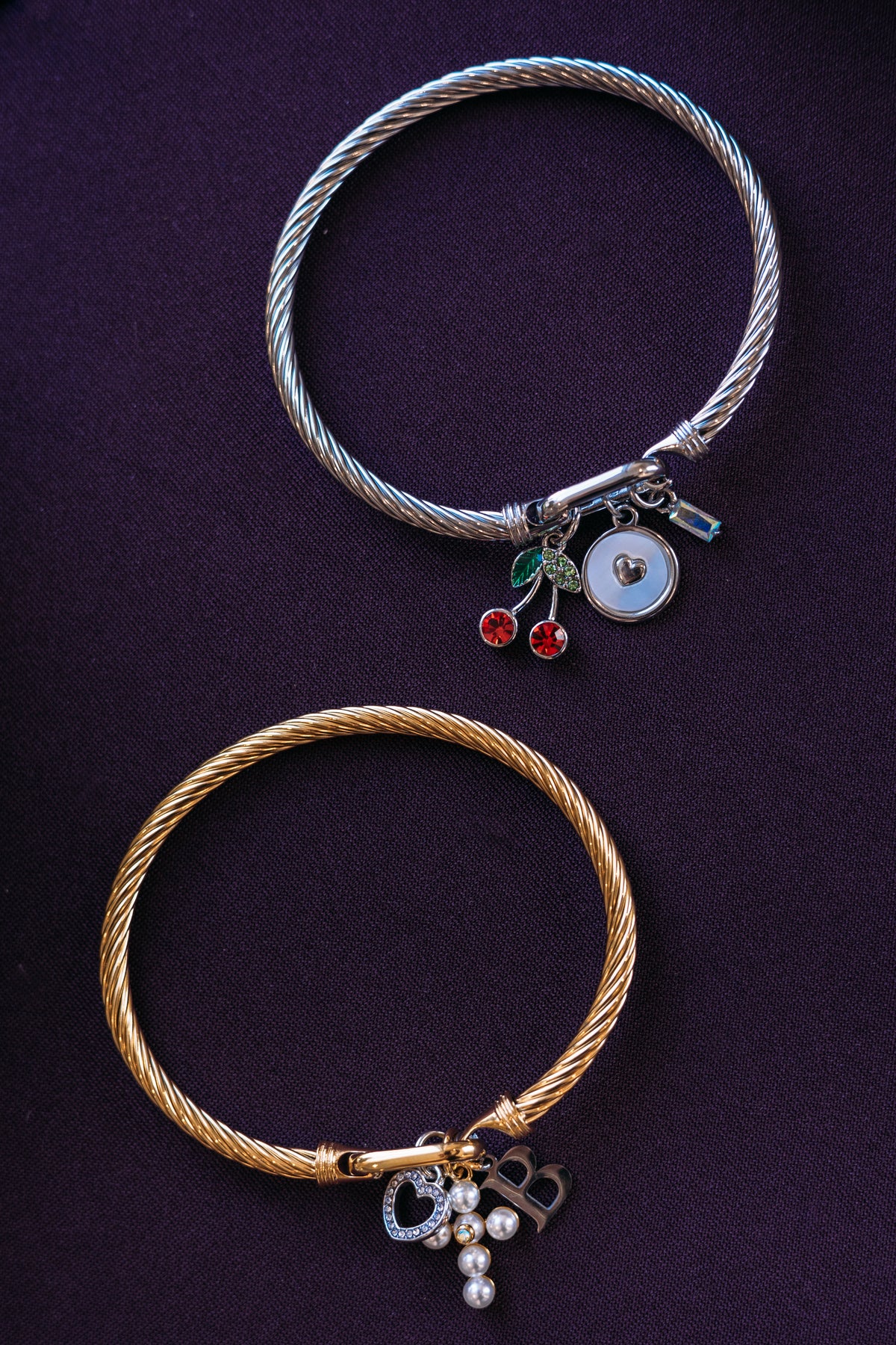 Two twisted cable-style bracelets on a dark purple background. The top bracelet is silver-tone with small charms including red cherries, green leaves, a round white charm, and a small blue bead.