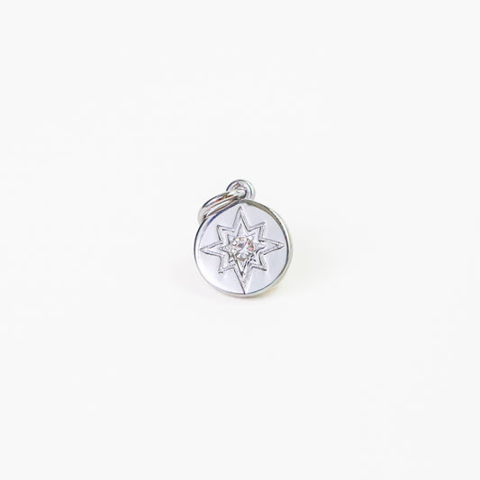 A silver-toned circular charm pendant featuring an engraved eight-pointed compass star design with a clear central gemstone, shown on a plain white background.