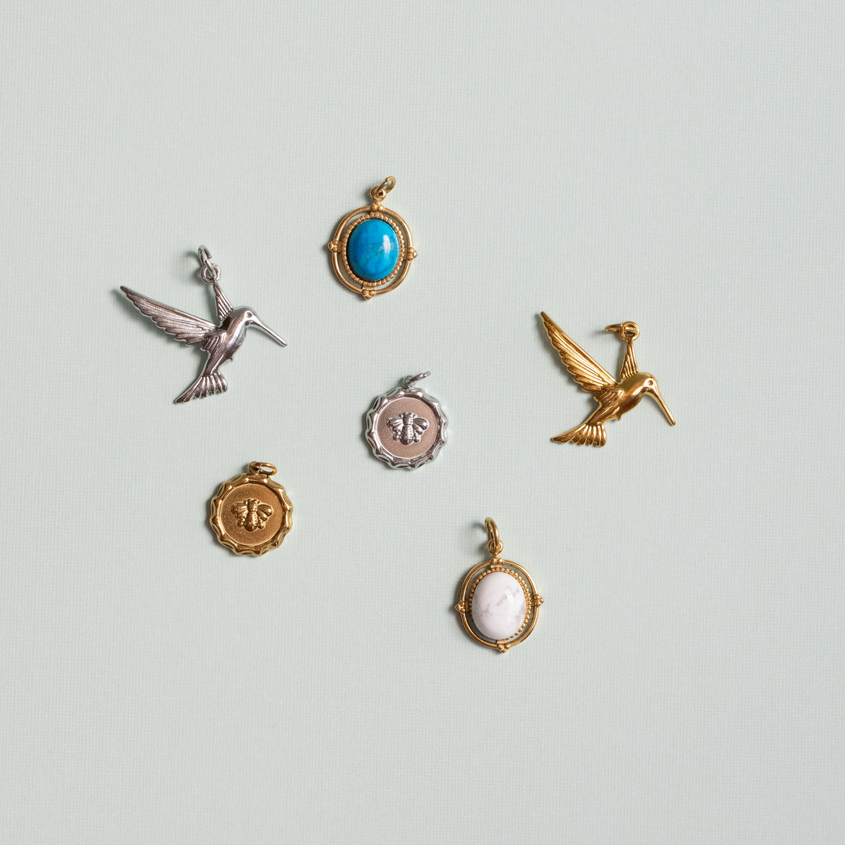 Six small jewelry charms or pendants arranged on a pale blue-gray background. The set includes two Hummingbird charms, in silver and gold,, one round blue stone pendant, and one round white stone pendant. The last pair of charms are two bee charms in silver and gold, the bee stamped on the charm like a letter wax. 