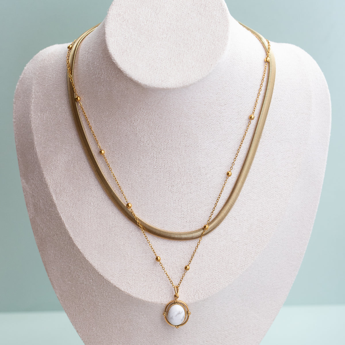 A double-layered gold necklace displayed on a cream-colored jewelry bust. The necklace features a textured snake chain layer and a delicate chain layer with small gold beads and a circular pendant containing a white Howlite Dot that lays flat off of the satellite necklace.