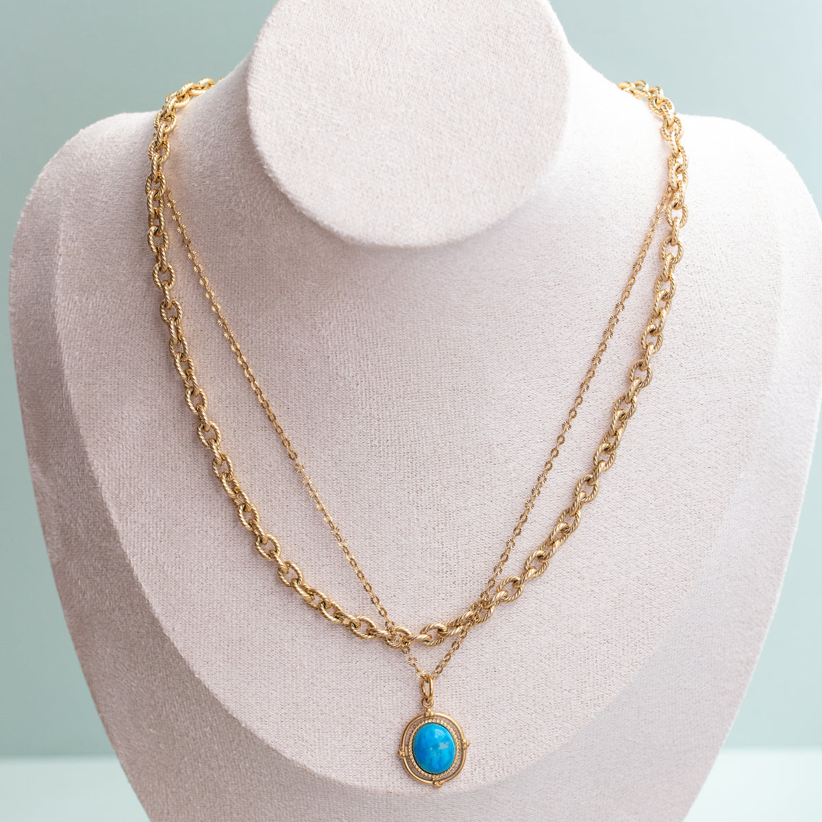 A gold double-strand necklace displayed on a light beige jewelry bust. The necklace features a textured chain and a delicate chain with a small oval turquoise -like pendant in a gold Foundation setting from Style Dots