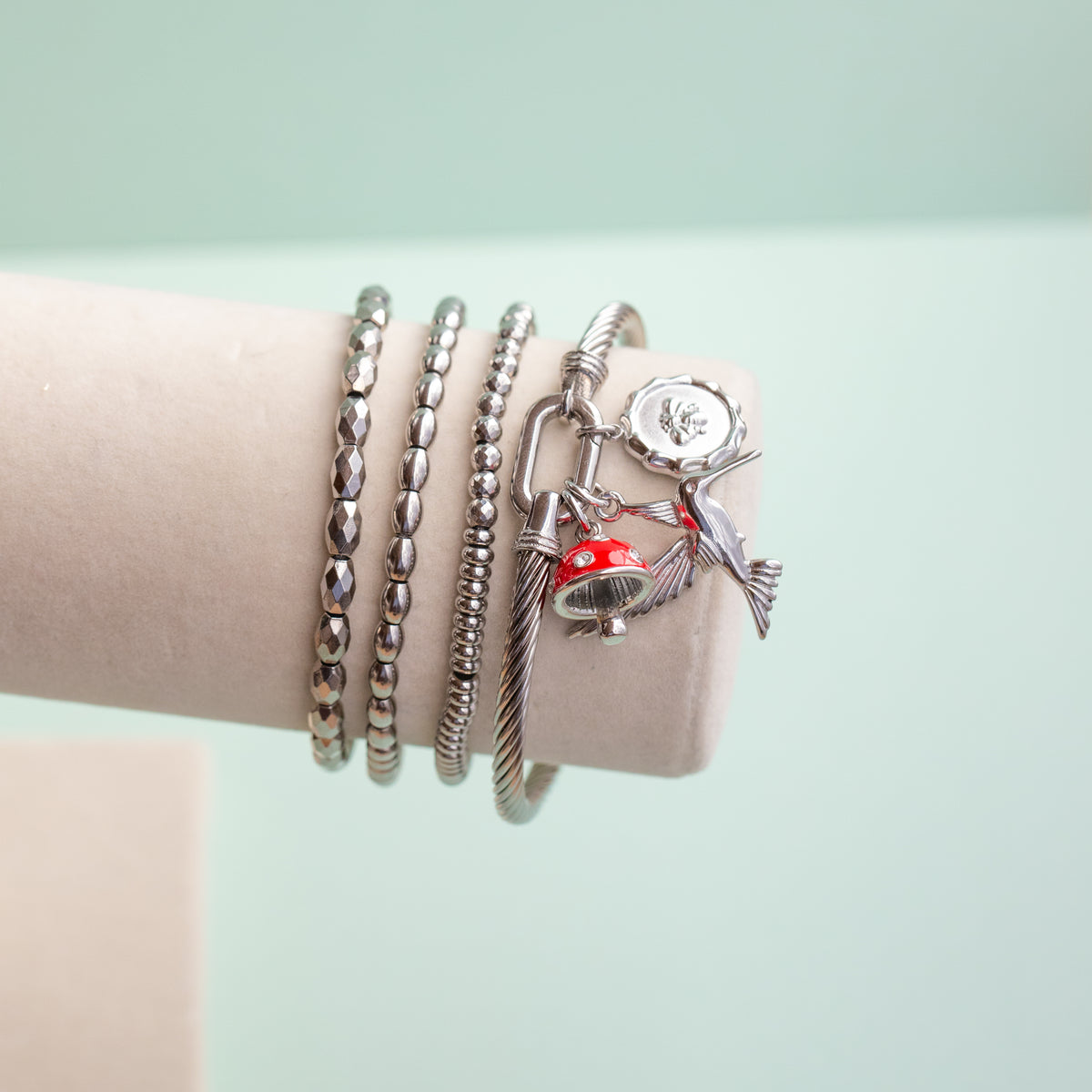 A set of four silver-toned bangles displayed on a light pink cylindrical jewelry stand. The bangles include a beaded bracelet, a textured twisted bracelet, a cable-knot style bracelet, and an open cuff bracelet featuring a silver hummingbird, a small red-and-white mushroom, and a round silver bee charm.