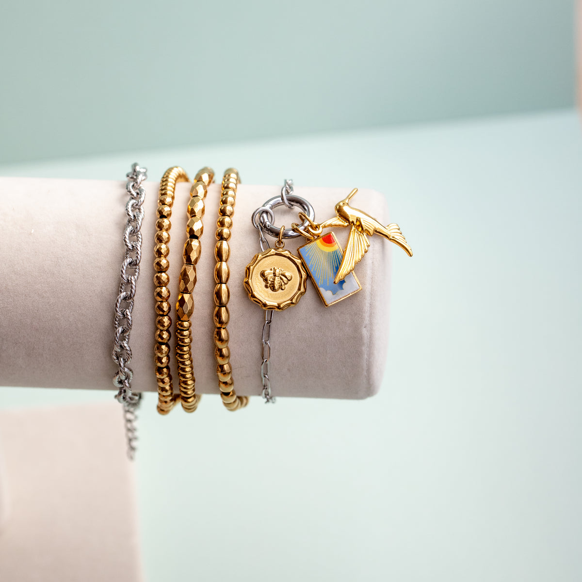 A set of gold and silver bracelets displayed on a light beige jewelry stand. The collection includes a silver chain bracelet, a gold beaded bracelet, a twisted gold bracelet, a silver charm keeper bracelet with a gold bee stamp charm, a small gold hummingbird charm, and a small blue and orange enamel charm. 