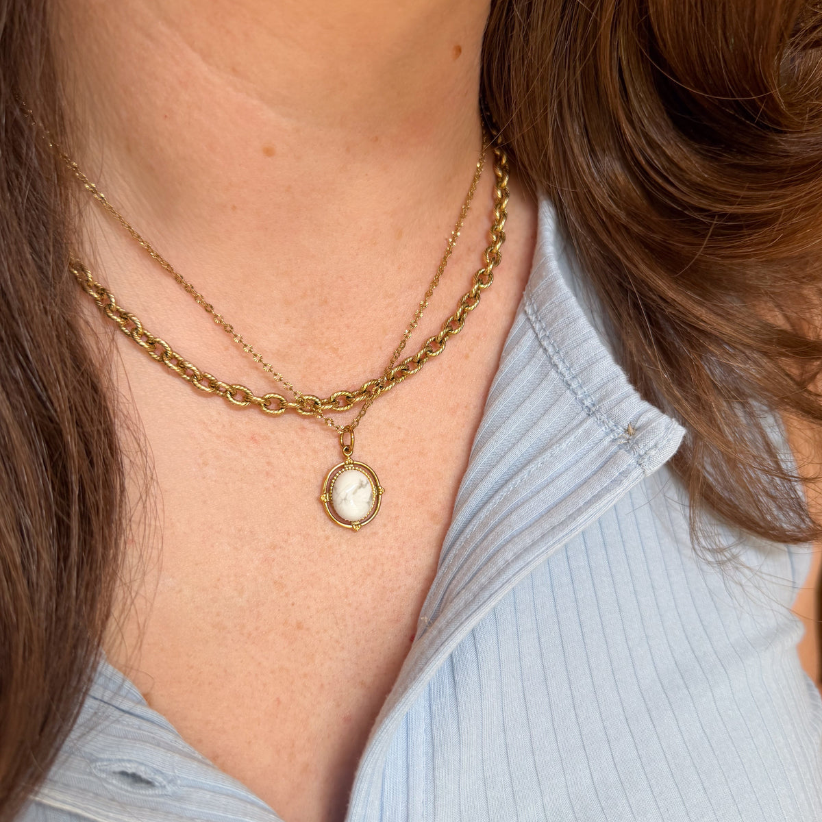 A close-up of a person wearing a double-layered gold necklace with a small Howlite Original Dot pendant, paired with a light blue ribbed top.