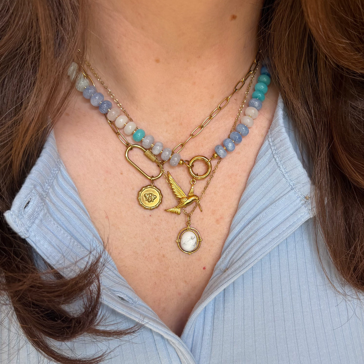 A close-up of a person wearing a layered necklace with gold-toned charms, including a bird and oval medallion, layered over a beaded strand with light blue, gray, and teal colored beads.