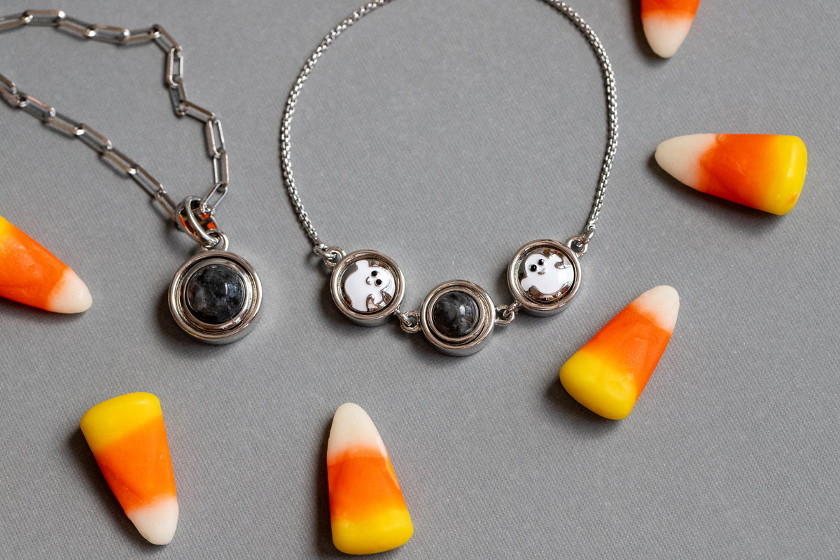 Two silver-toned jewelry pieces on a gray background, styled with candy corn pieces. One is a paperclip chain necklace with a round dark stone pendant, and the other is a delicate chain bracelet with three circular charms, including one with a white ghost design and two with black flashstones.