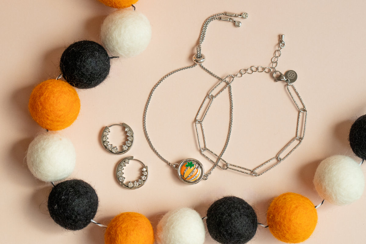 Silver-toned jewelry set including a paperclip chain bracelet, a bolero bracelet with a round pumpkin charm, matching dot, and a felt beaded garland with orange, white, and black felt balls, arranged on a pale peach background. 2026 Halloween. 2 Inside Crystal Hoop Earrings are paired with the flat bracelet stack. 