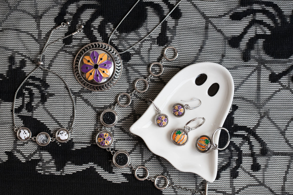 Halloween-themed jewelry set featuring a silver-toned necklace and matching earrings with colorful floral and pumpkin charms, displayed on a black spider-patterned fabric next to a white ghost-shaped dish with a lace background. 