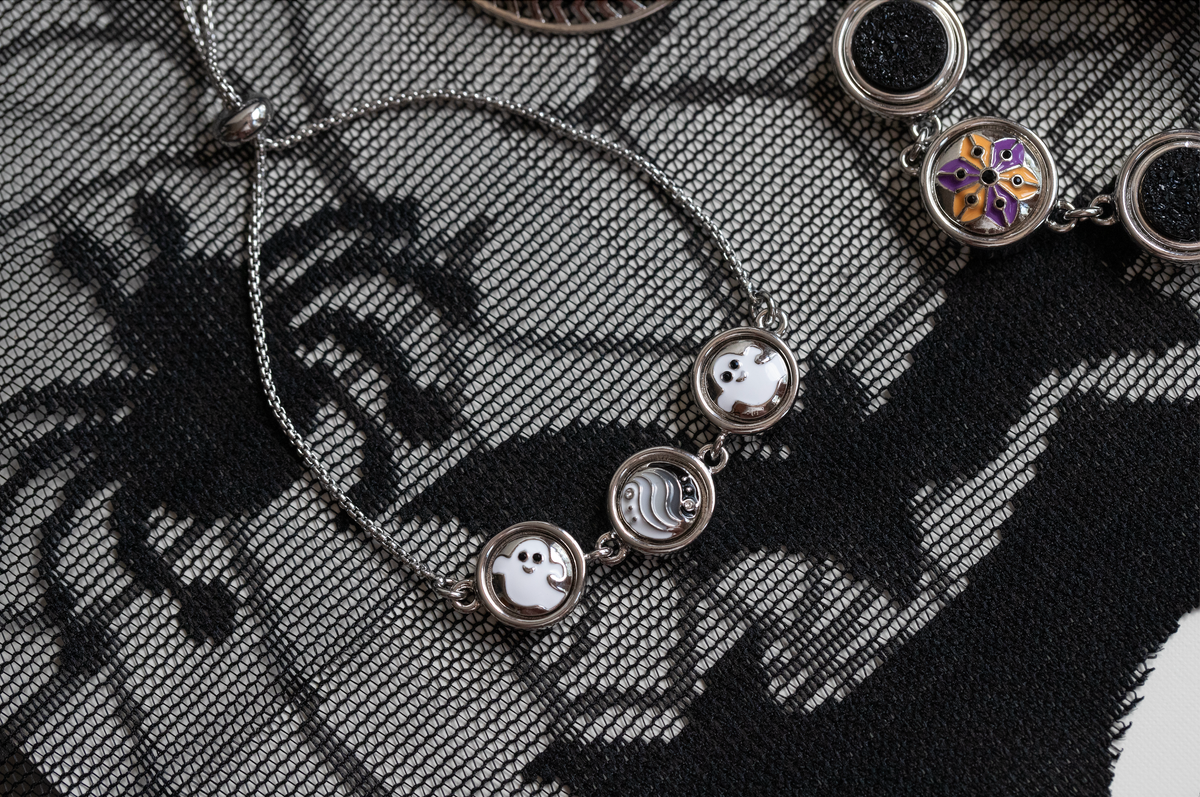 A close-up of a silver-toned necklace with three circular charms featuring ghost and wave designs, laid on a black and gray lace fabric background for Halloween 2026