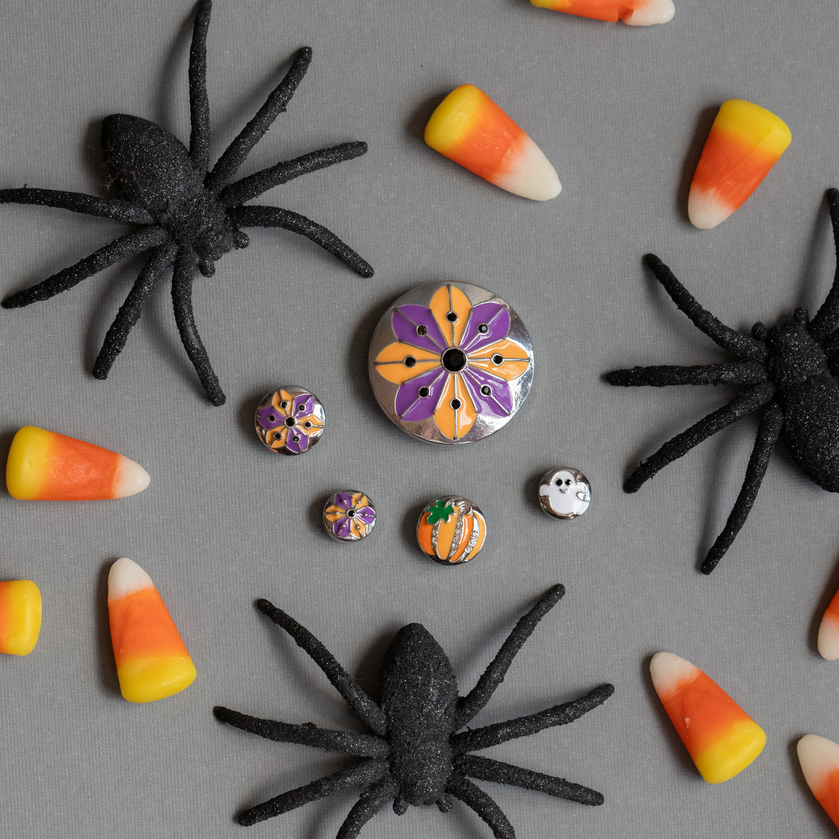 A flat-lay of Halloween-themed jewelry findings on a gray background, including a central round Dot with a purple, orange, and yellow floral pattern, two smaller matching patterned charms, a small white ghost and pumpkin charm, with glittery black spider decorations surrounded by candy corn props. Launched for Halloween 2026