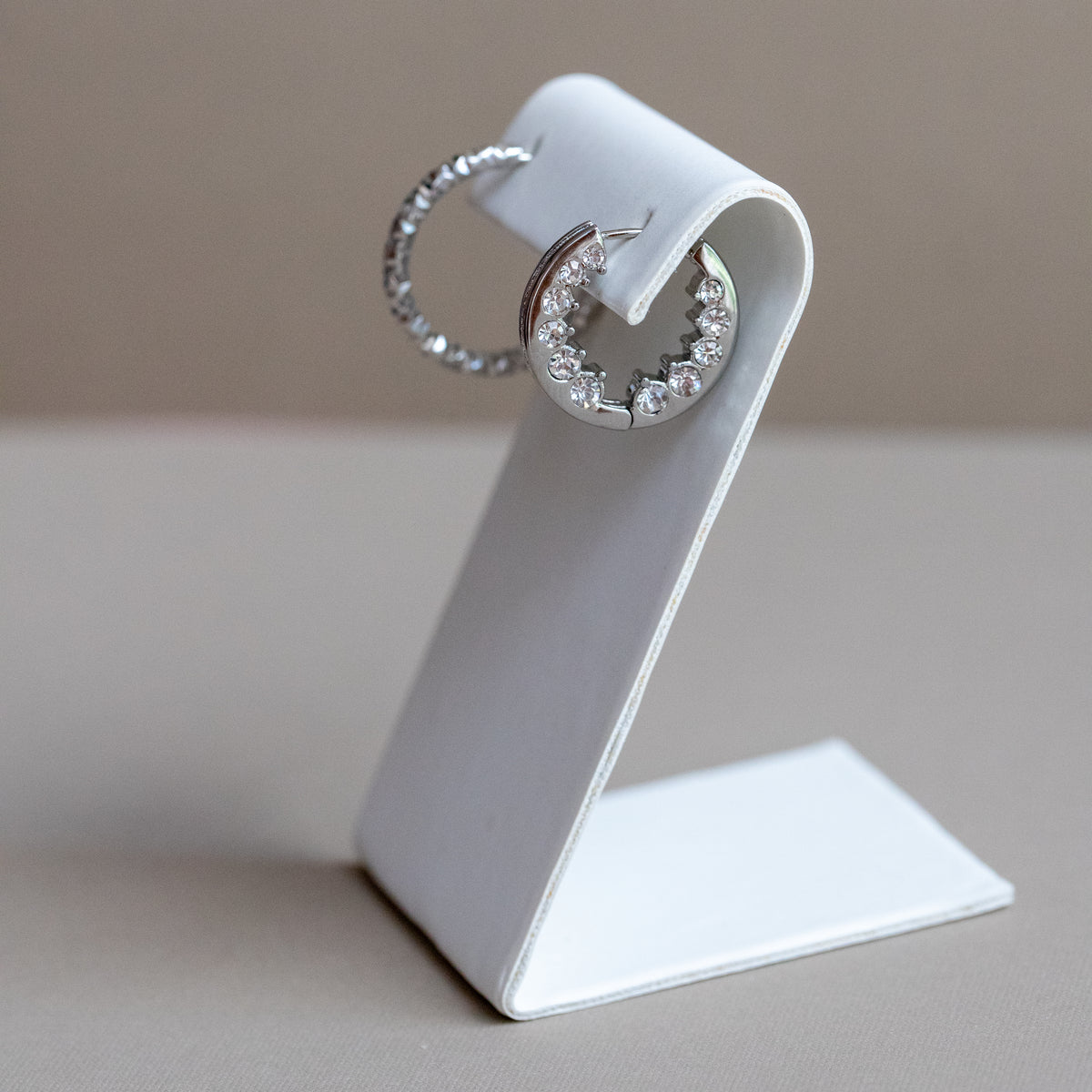 A pair of silver crescent-shaped hoop earrings with small clear stones, displayed on a white earring stand against a warm beige background.