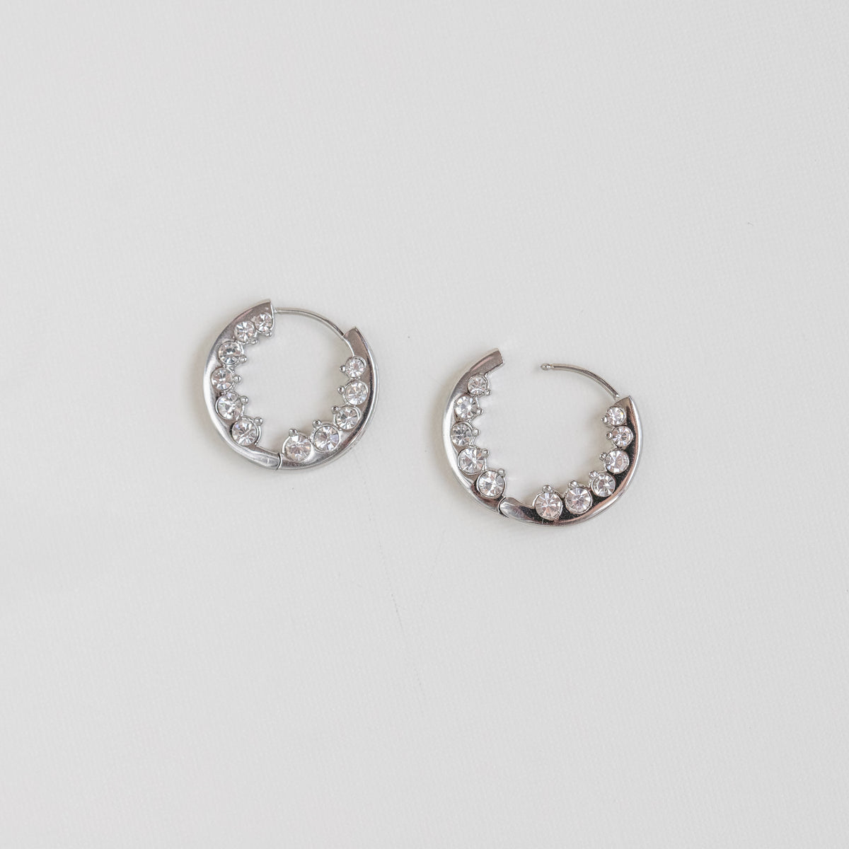 A pair of silver crescent-shaped hoop earrings embellished with clear rhinestones, displayed on a clean white background.