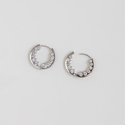 A pair of silver crescent-shaped hoop earrings embellished with clear rhinestones, displayed on a clean white background.