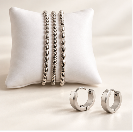 A jewelry set featuring three silver beaded bracelets and a pair of silver hoop earrings, displayed on a white cushion and a light beige surface.