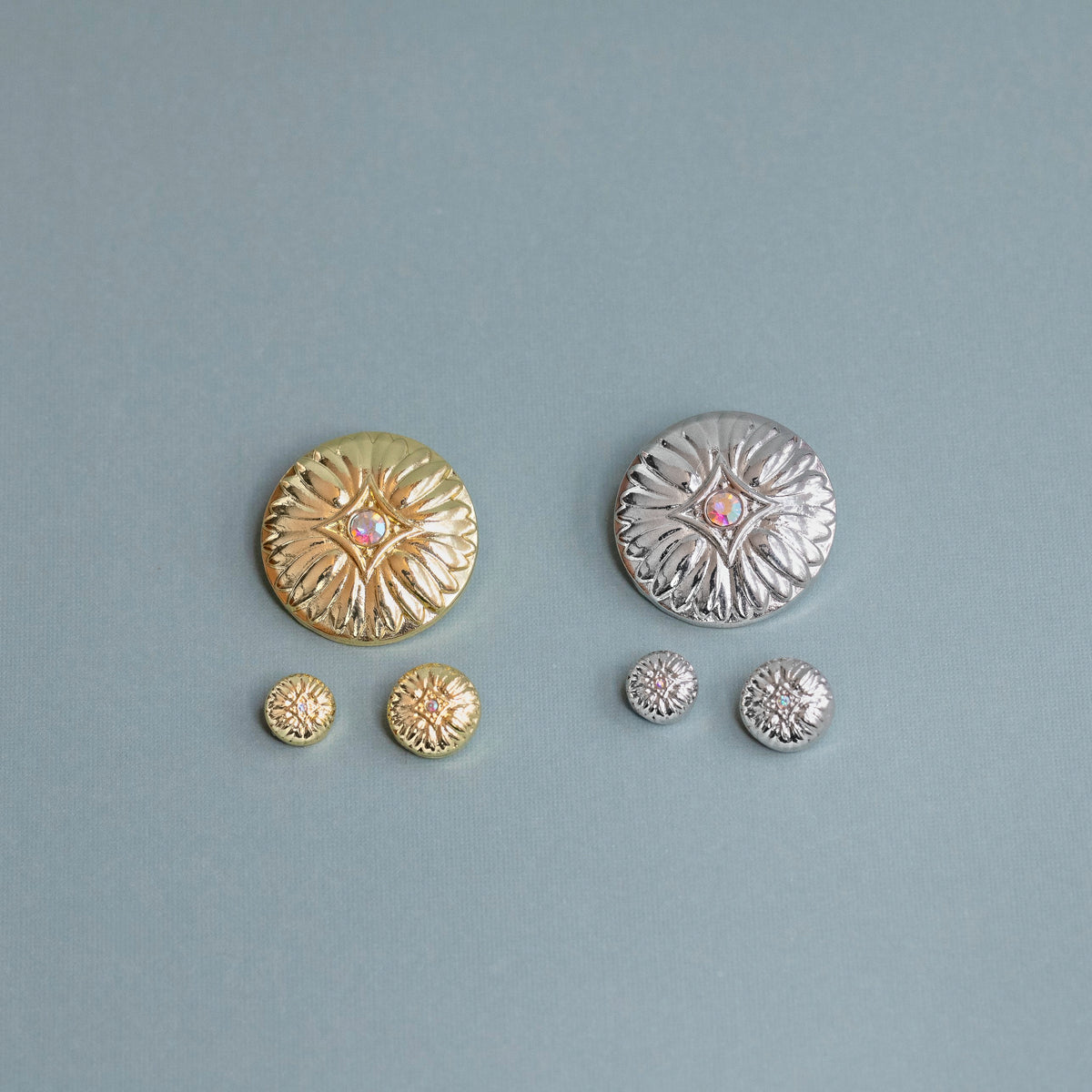 Two pairs of round textured stud earrings on a dusty blue background. One pair is gold-toned with a small pink center stone, and the other pair is silver-toned with a small pink center stone.