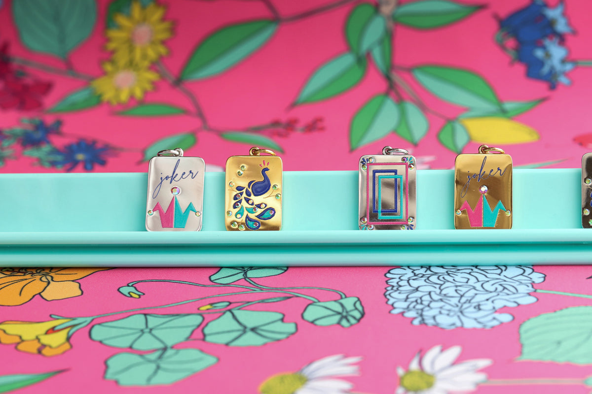 A close-up of four colorful rectangular jewelry pendants displayed on a turquoise tray, against a bright pink Mahjong floral mat. The pendants feature decorative illustrations and script lettering, including a peacock design and gold-toned lettering. The charms are in silver and gold.  The jewelry collection is inspired by Eastern Asian Mahjong tiles.