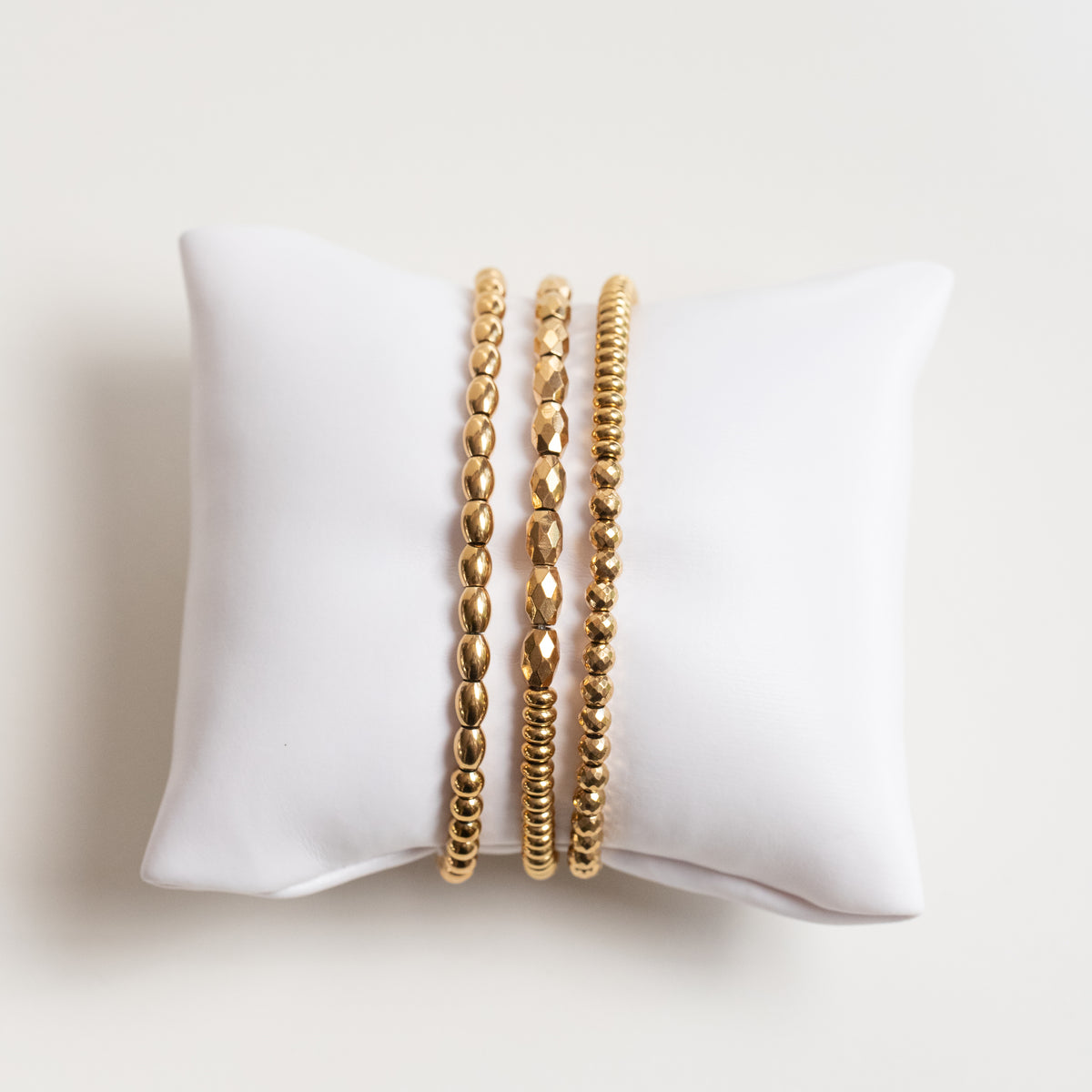Three gold beaded stretch bracelets displayed on a white square pillow against a light beige background.
