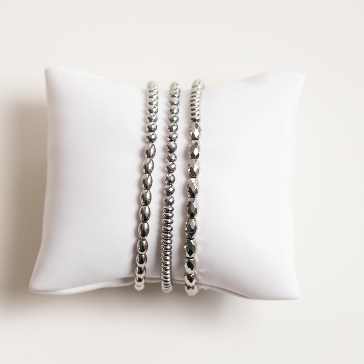 Three silver beaded stretch bracelets displayed on a white fabric jewelry pillow against a light beige background.
