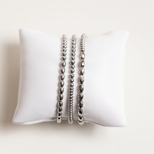 Three silver beaded stretch bracelets displayed on a white fabric jewelry pillow against a light beige background.