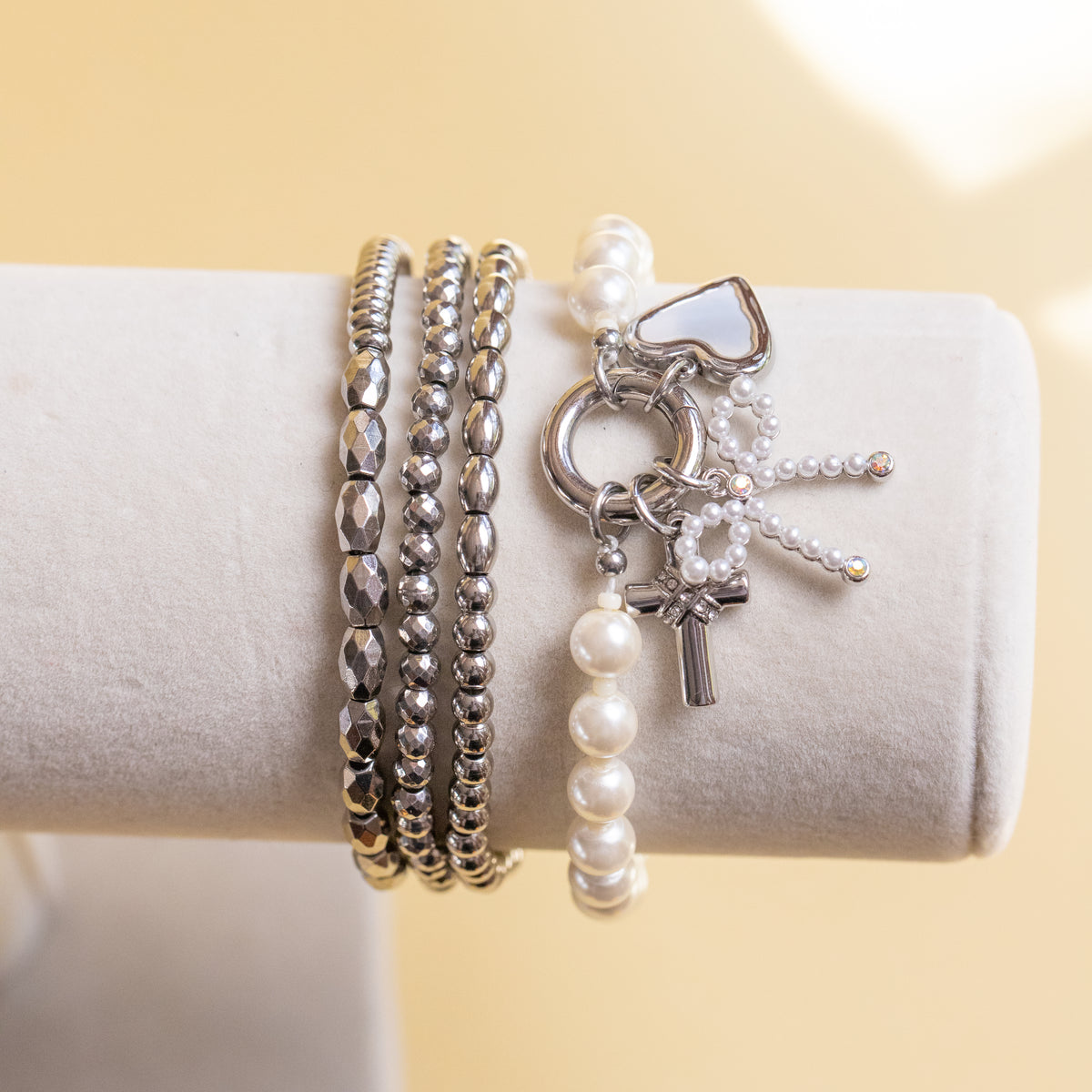 A set of silver and pearl bracelets displayed on a beige cylindrical jewelry stand against a soft beige background. The bracelets include textured silver beaded bands, a silver charm bracelet with a heart-shaped stone, a cross charm, and a pearl-and-silver key-style charm.