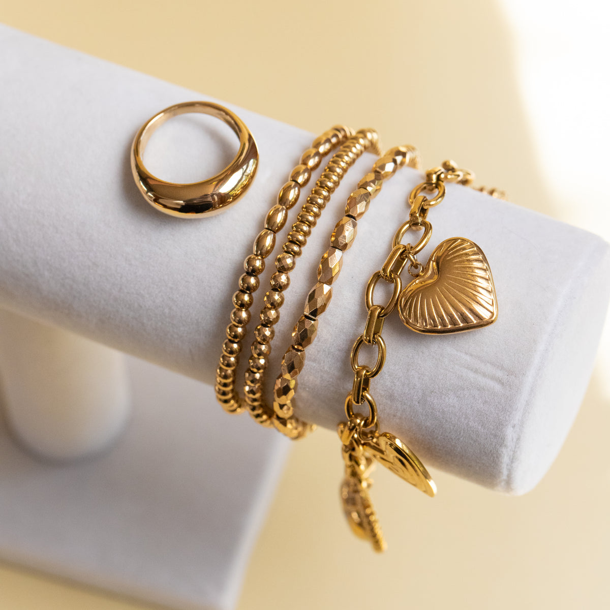 A set of gold-toned jewelry displayed on a white cylindrical stand, including a thick textured ring, three beaded and chain bracelets, and a heart-shaped charm.
