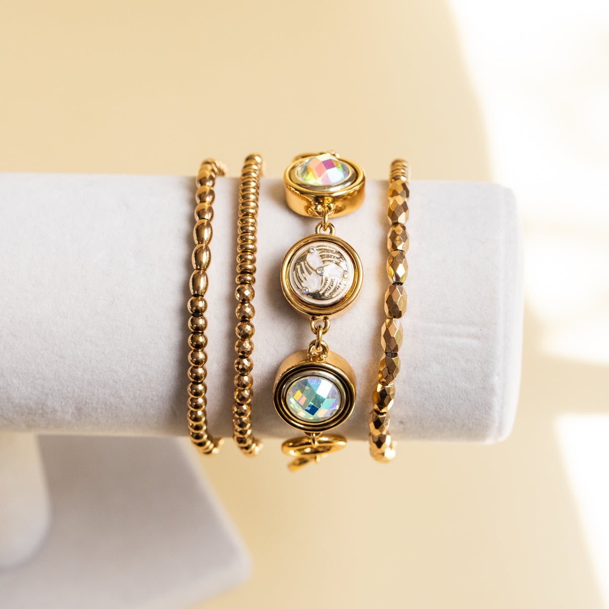 Four gold-toned bracelets displayed on a white jewelry stand against a warm beige background. The set includes a beaded chain bracelet, a textured link bracelet, a round bezel charm bracelet with a silver-toned globe medallion, and a round bezel charm bracelet with an iridescent blue-green stone.
