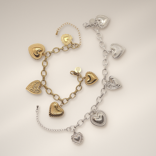 Two heart-themed charm bracelets displayed on a light beige background. One bracelet is gold with textured and polished heart charms, and the other is silver with matching heart charms. 