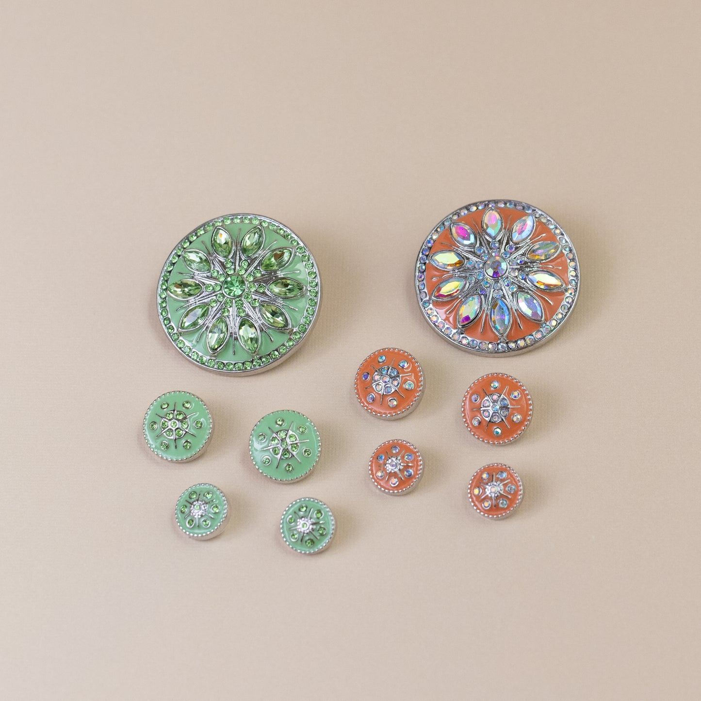 A set of decorative round dots with rhinestone floral designs, including two larger mint green and two larger orange buttons, plus several smaller matching buttons, arranged on a solid beige background.