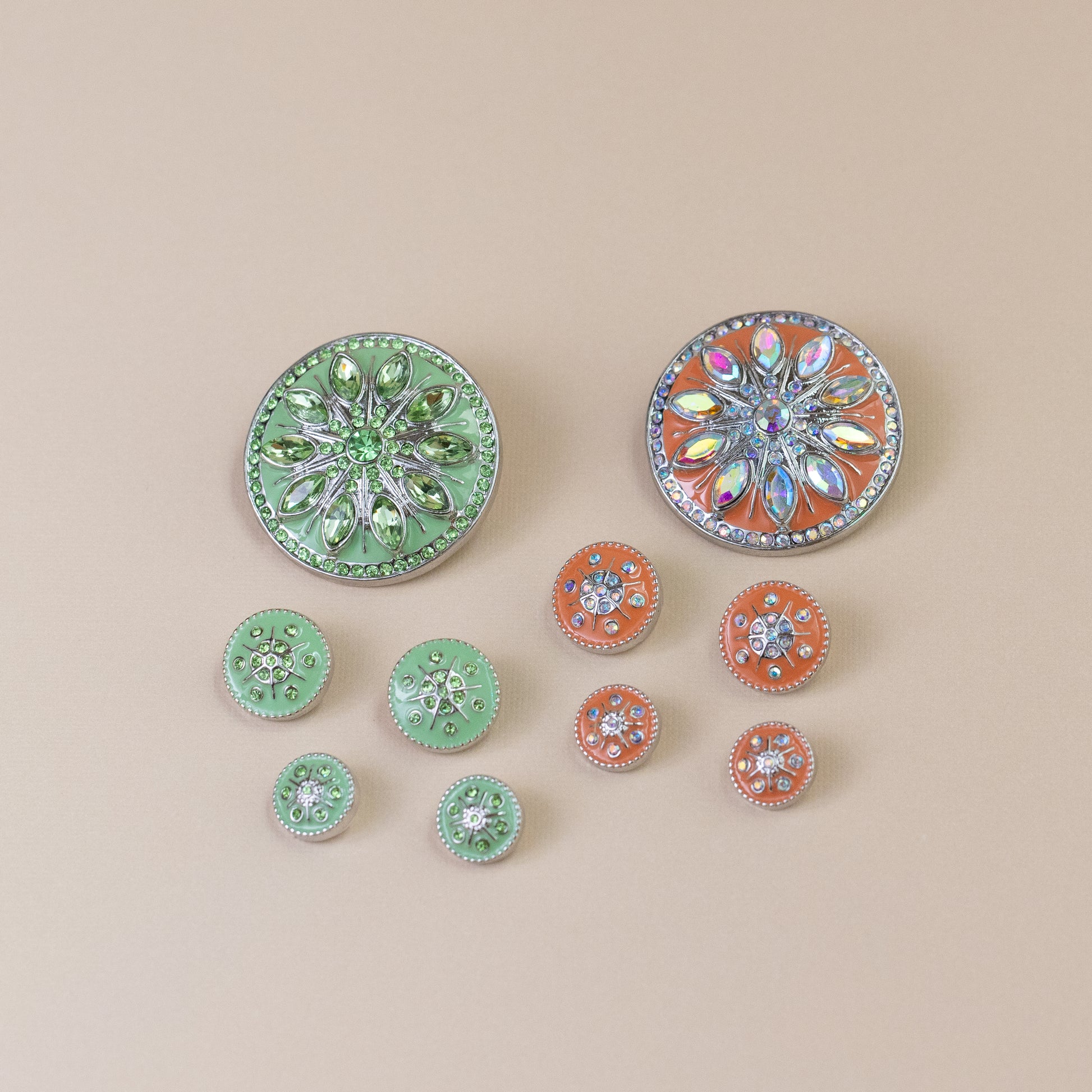 A set of decorative round dots with rhinestone floral designs, including two larger mint green and two larger orange buttons, plus several smaller matching buttons, arranged on a solid beige background.