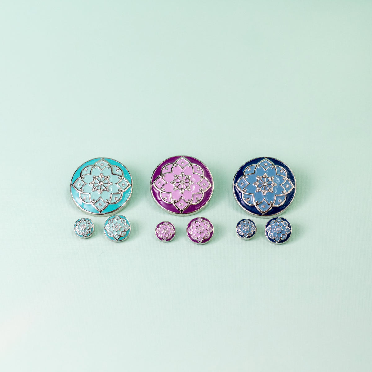 Three pairs of round floral enamel jewelry pieces arranged on a pale mint background, with larger interchangeable Dots above smaller matching Dot sizes. The top row features light aqua, magenta, and periwinkle blue designs with floral detailing and dark outlines; the bottom row shows matching smaller pieces in the set