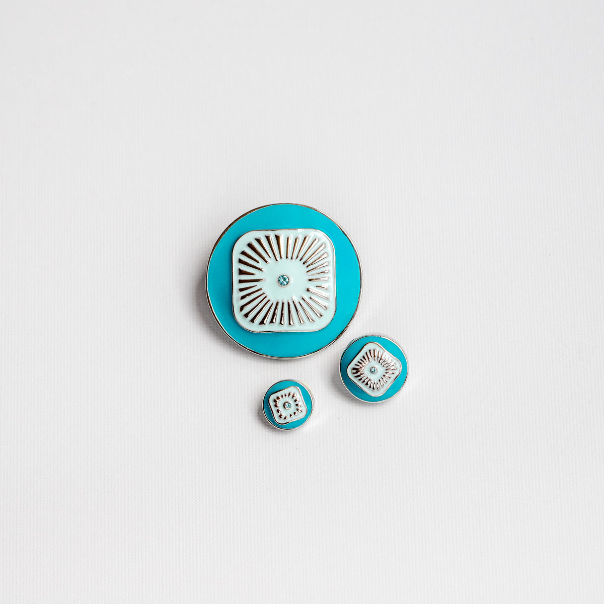 A set of 3 round turquoise and light blue Dots in all available sizes, centered in the middle of a white textured background. A small matching blue crystal is set inside a sunburst design on all 3 Dots.