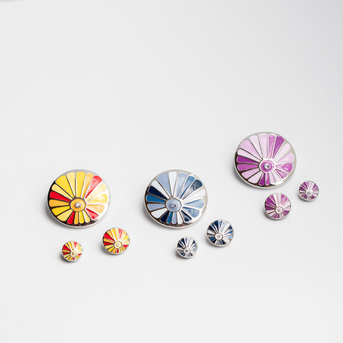 A bundle of colorful circular enamel-style pins or buttons with fan sunburst patterns, arranged on a white background. The designs include yellow and red, blue and white, and purple and silver versions, with all of the smaller matching Dots below.