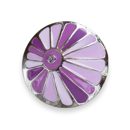 A circular enamel button featuring a sunburst pattern in shades of purple and lavender, outlined with a silver-tone metal border and a small clear rhinestone in the center, shown on a plain white background.