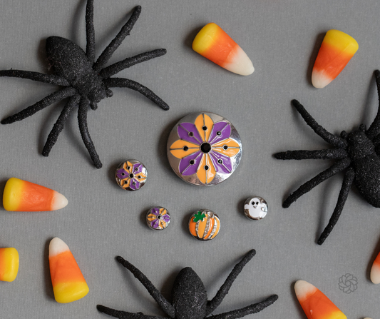 A styled flat-lay of small round craft beads on a gray background, surrounded by glittery, black spider decorations, candy corn, and a small pumpkin. The central Statement Dot features a purple and orange floral mandala pattern on a silver backing, with smaller coordinating Dots nearby in the Original and Dotlet Styles. This product drop is from September 2026. 