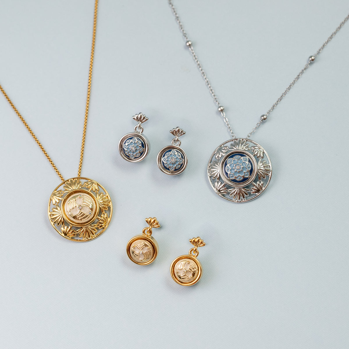 A jewelry set featuring two round pendant necklaces and four matching earrings, displayed on a light gray background. One necklace is gold-toned with a circular medallion pendant, the other is silver-toned with a matching medallion pendant, and the chains are a box chain and satellite chain respectively.